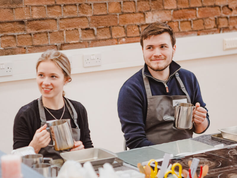 Two people sitting in grey aprons holding stainless steel jugs in a candle making class